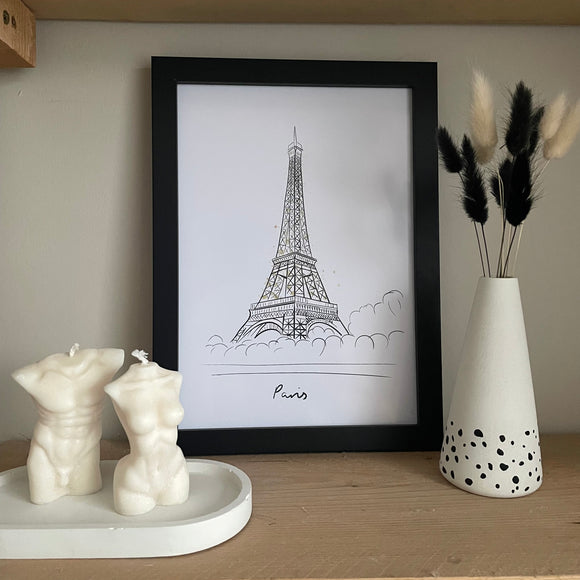 Travel Print of the Eiffel Tower in Paris with sparkling lights. A4 in a black frame. Dalmatian print concrete vase with bunny tails to the right and oval tray with body candles to the left