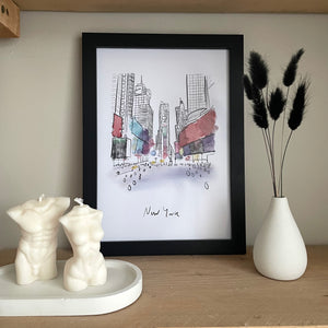 A4 New York Wall Art Print. Busy Times Square with splashes of colour. A4 in black frame surrounded by concrete vase and black bunny tails and a concrete oval tray with body candles